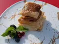 Delicioso postre de milhojas de crema, con capas crujientes y relleno dulce, acompañado de frutos rojos y menta fresca.