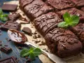 Deliciosos postres de chocolate, brownies cubiertos con glaseado de chocolate y decorados con menta fresca. Trozos de chocolate y una cuchara con chocolate derretido completan la escena.