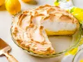 Tarta de limón con merengue dorado y esponjoso, lista para servir. Dos limones frescos y un paño con estampado de limones completan la escena.