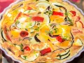 Quiche de verduras con hojaldre, repleta de pimientos, calabacín y cebolla morada, adornada con hierbas frescas.