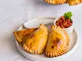 Tres empanadillas rellenas doradas y crujientes, servidas con una pequeña taza de salsa de tomate y una hoja de perejil.