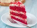 Rebanada de torta red velvet con glaseado blanco y migas rojas, servida en plato blanco con tenedor.