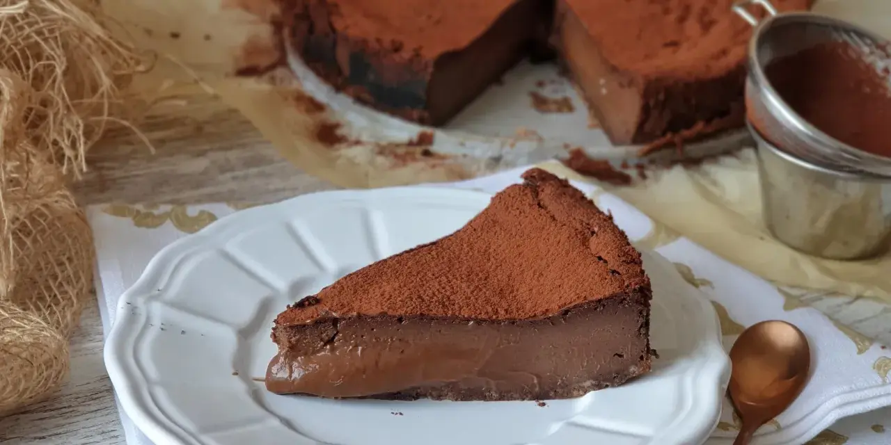 Una rebanada de torta de chocolate, cubierta de cacao en polvo, reposa en un plato blanco. El postre se ve cremoso y delicioso.