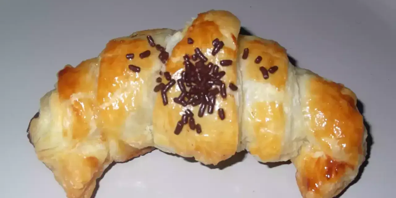 Croissant de chocolate recién horneado, dorado y brillante, espolvoreado con virutas de chocolate.