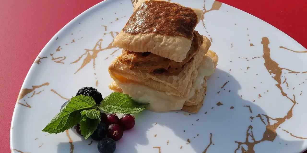 Delicioso postre de milhojas de crema, con capas crujientes y relleno dulce, acompañado de frutos rojos y menta fresca.
