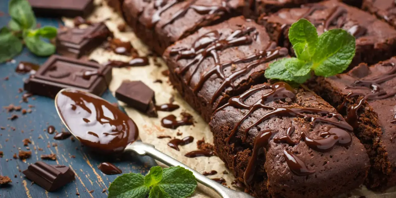 Deliciosos postres de chocolate, brownies cubiertos con glaseado de chocolate y decorados con menta fresca. Trozos de chocolate y una cuchara con chocolate derretido completan la escena.