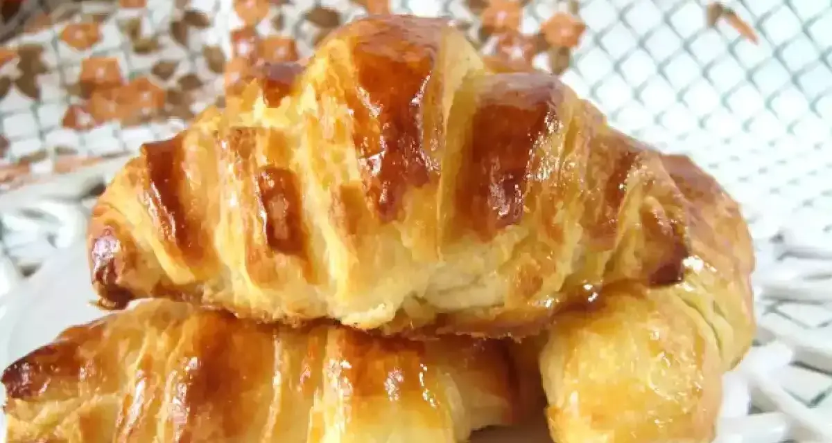 Dos croissants caseros dorados y brillantes, apilados en un plato blanco.