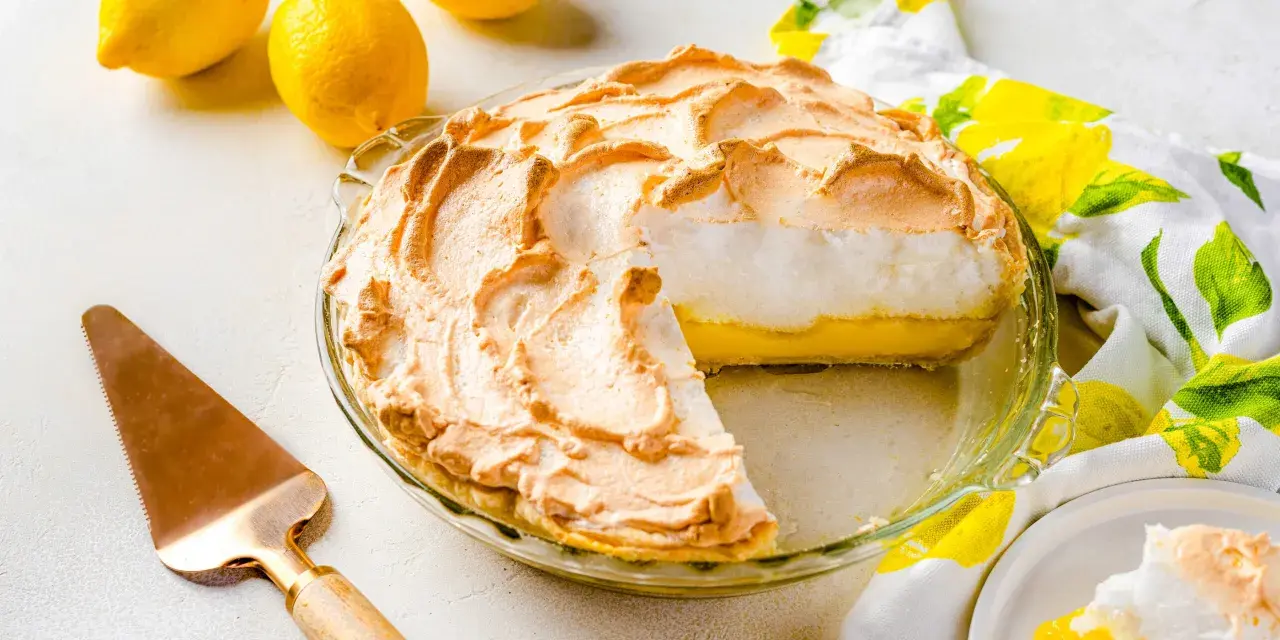 Tarta de limón con merengue dorado y esponjoso, lista para servir. Dos limones frescos y un paño con estampado de limones completan la escena.
