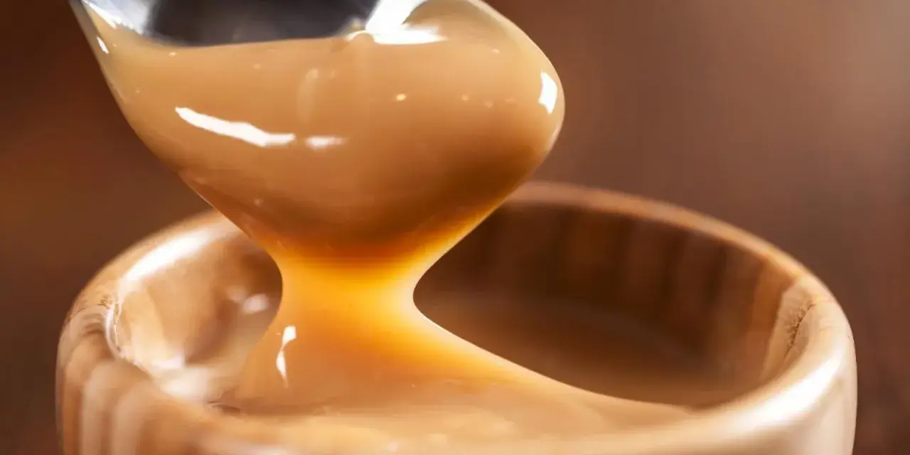 Cuchara vertiendo manjar blanco cremoso en un cuenco de madera.