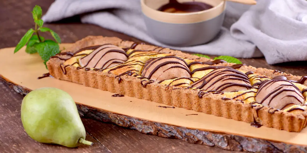 Tarta rectangular con peras y chocolate, decorada con menta. Una pera fresca reposa a un lado.