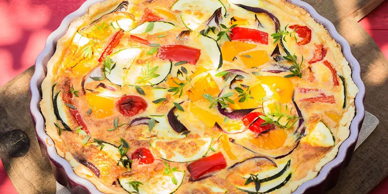 Quiche de verduras con hojaldre, repleta de pimientos, calabacín y cebolla morada, adornada con hierbas frescas.