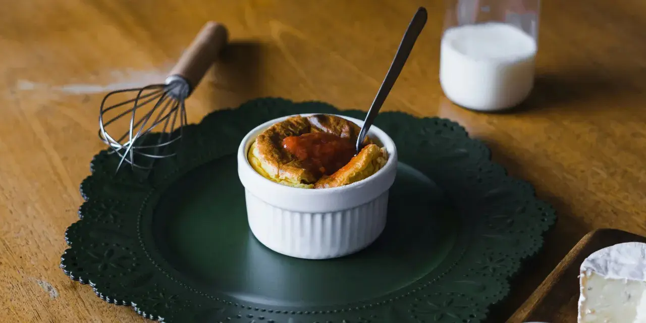 Un esponjoso soufflé con salsa de tomate, listo para disfrutar. Al lado, un batidor y un vaso de leche, ingredientes clave para crear que es un soufflé.