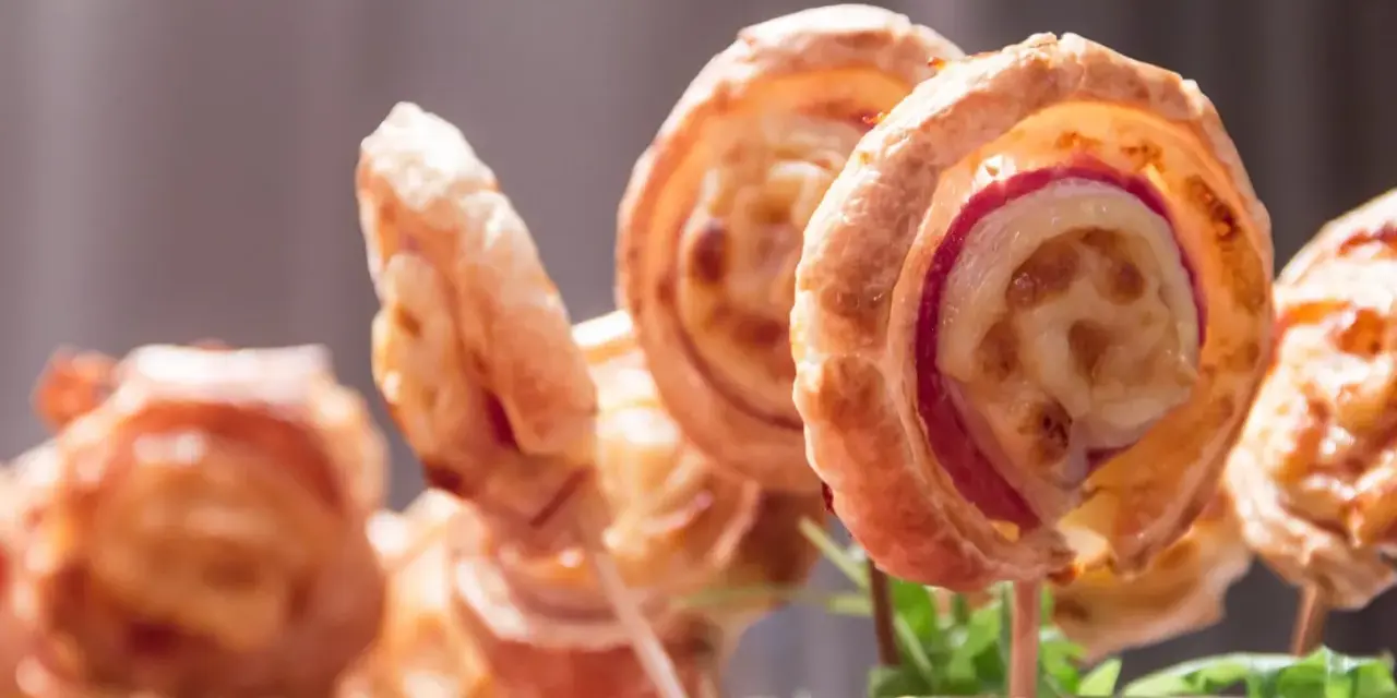 Deliciosas piruletas de hojaldre rellenas de queso y jamón, presentadas en un vaso con hojas verdes.