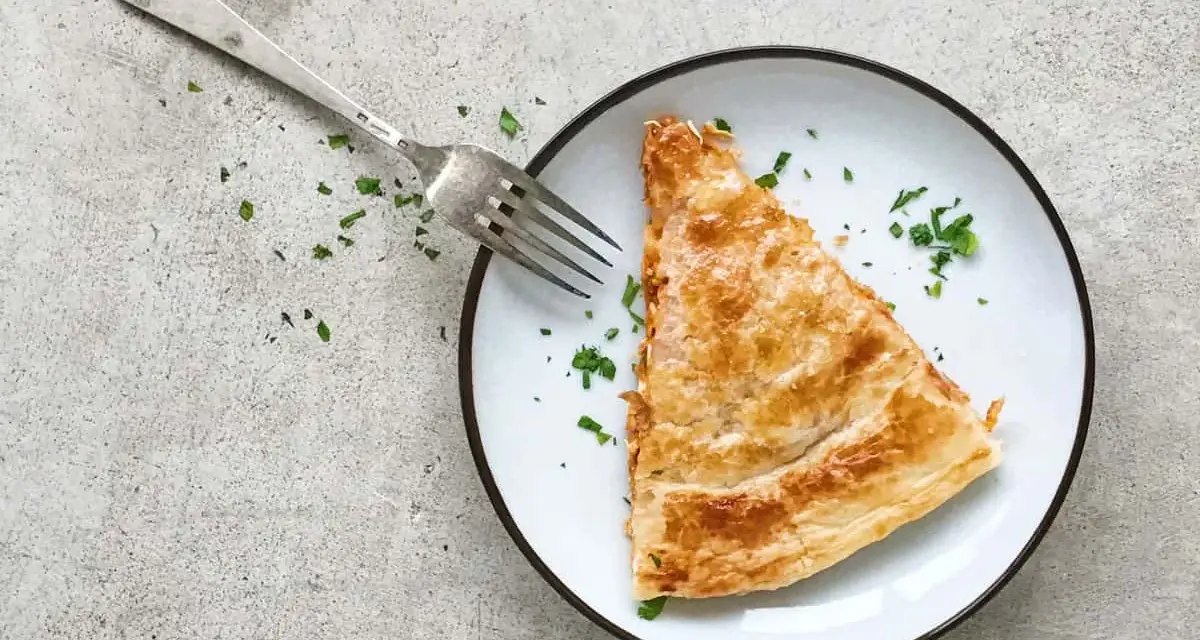 Una porción dorada de empanada de atún, lista para ser disfrutada con un tenedor plateado y espolvoreada con perejil fresco.