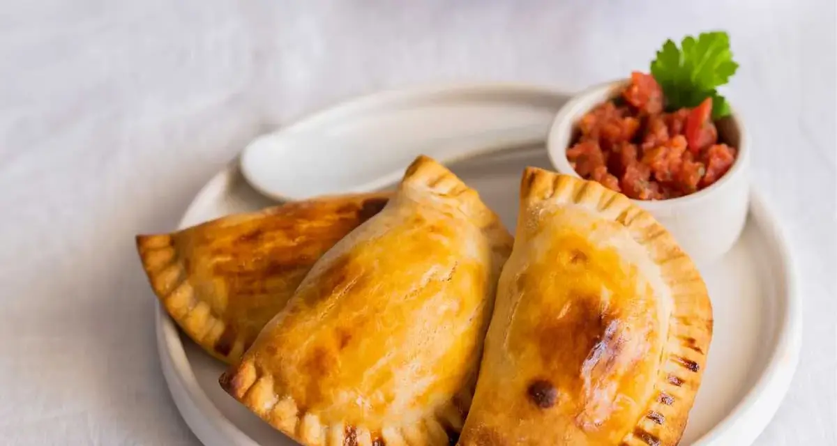 Tres empanadillas rellenas doradas y crujientes, servidas con una pequeña taza de salsa de tomate y una hoja de perejil.