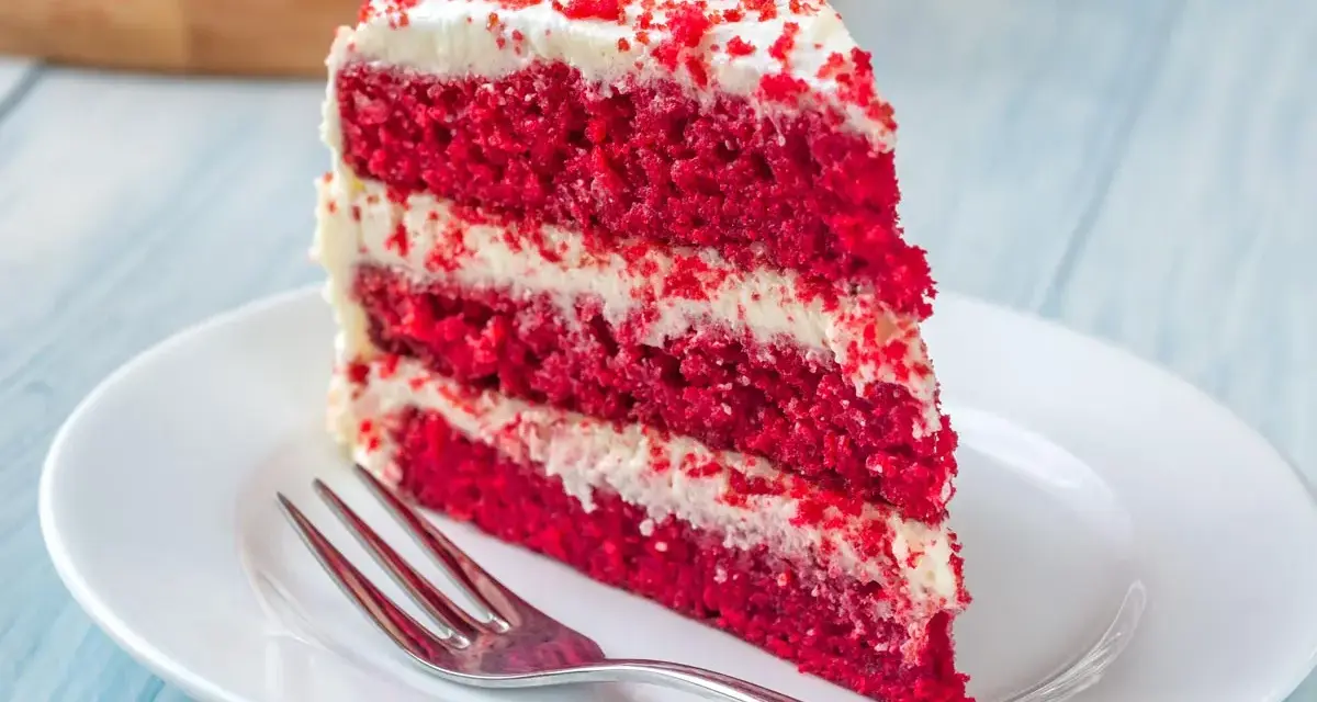 Rebanada de torta red velvet con glaseado blanco y migas rojas, servida en plato blanco con tenedor.
