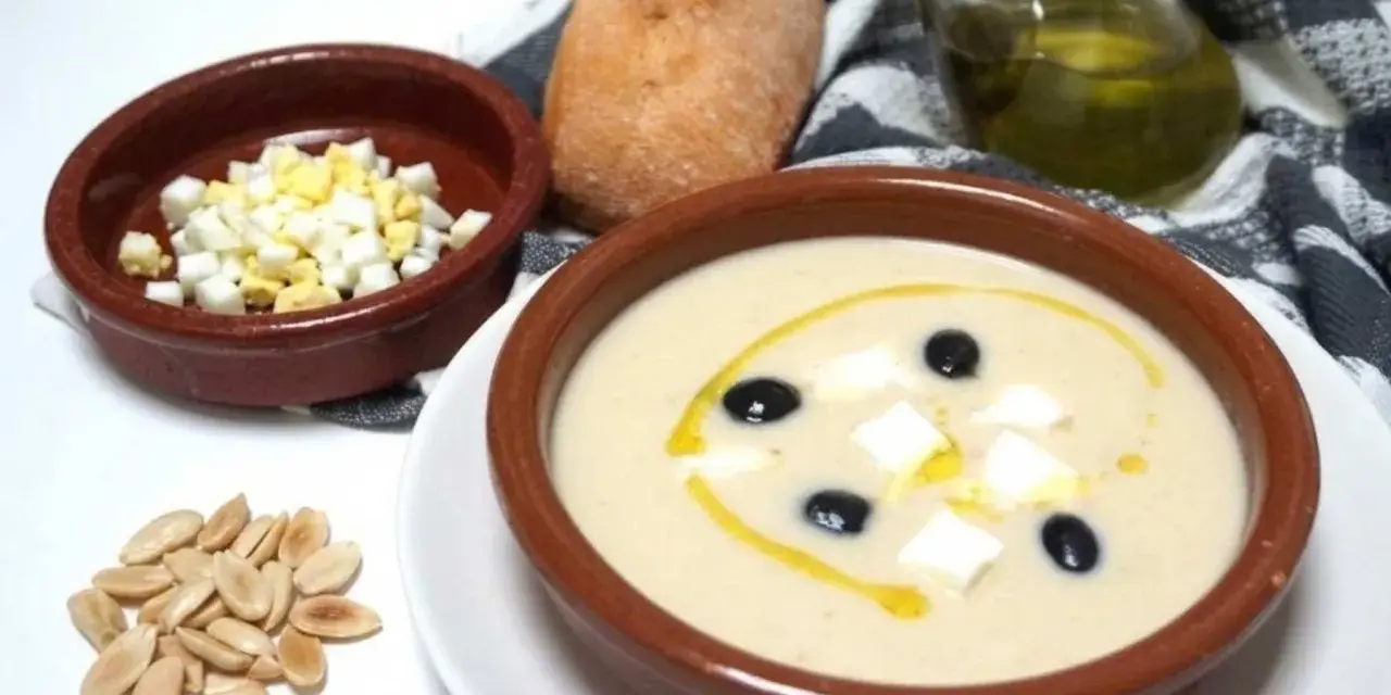 Un cuenco de mazamorra cordobesa con aceitunas, huevo duro y un chorrito de aceite. Acompaña un panecillo y almendras.