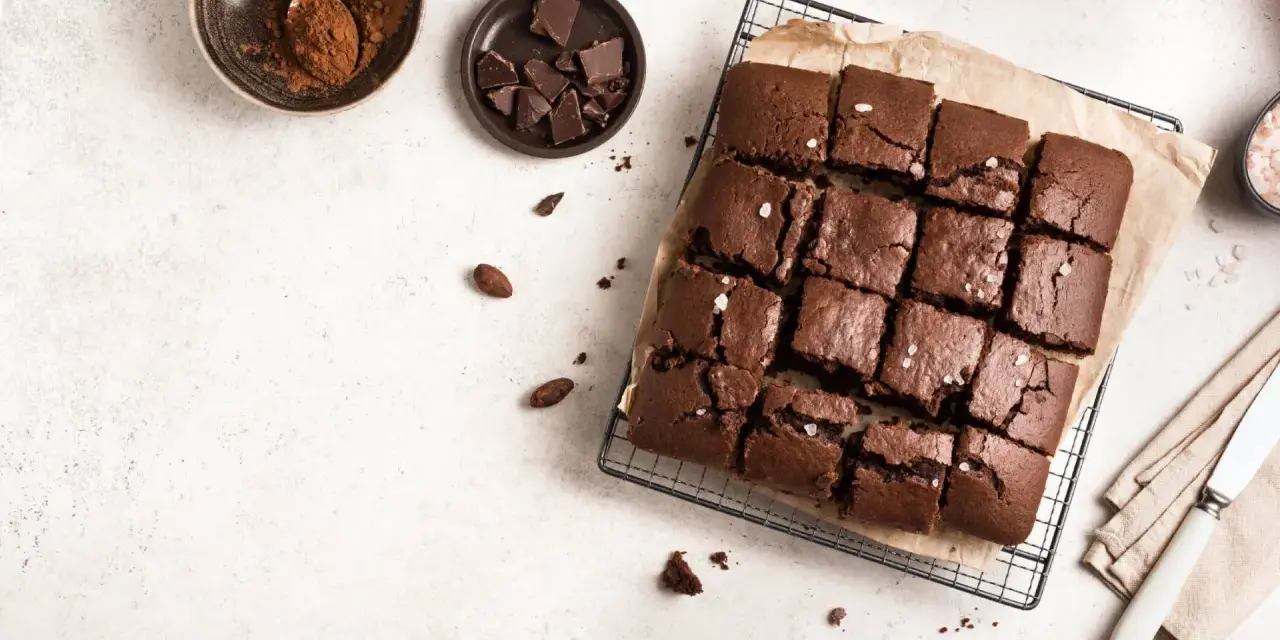 Delicioso brownie sin horno, cortado en cuadrados y espolvoreado con sal marina. Ingredientes como cacao y chocolate en trozos acompañan esta tentación.