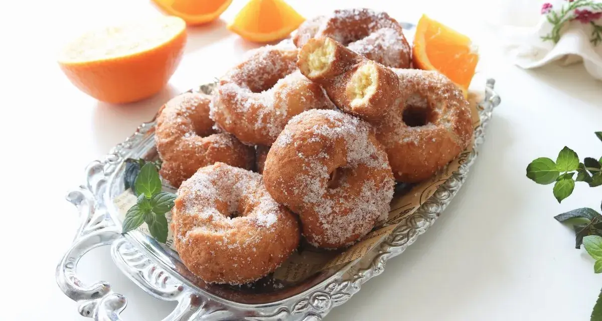 Rosquillas de naranja recién hechas, espolvoreadas con azúcar, junto a gajos de naranja fresca en una bandeja plateada.