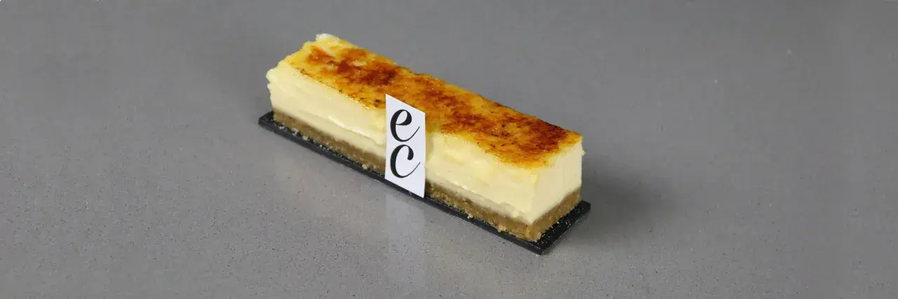 Un trozo de cheesecake creme brulee con base crujiente y cubierta caramelizada, adornado con un distintivo.