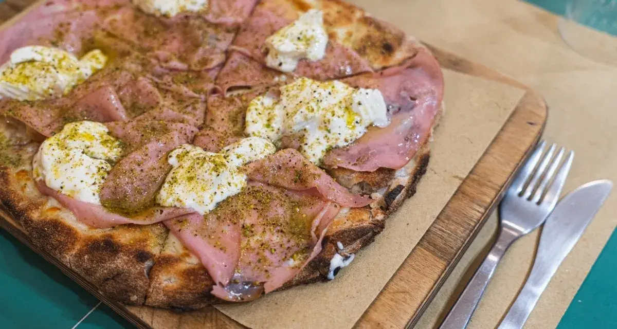 Pizza con mortadela y burrata cremosa, espolvoreada con hierbas verdes y servida en tabla de madera.