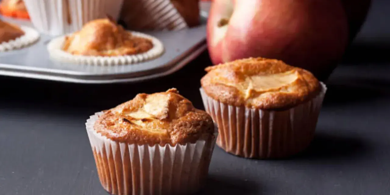 Dos deliciosos muffins de manzana recién horneados, con una manzana roja al fondo.