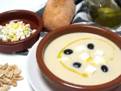 Un cuenco de mazamorra cordobesa con aceitunas, huevo duro y un chorrito de aceite. Acompaña un panecillo y almendras.