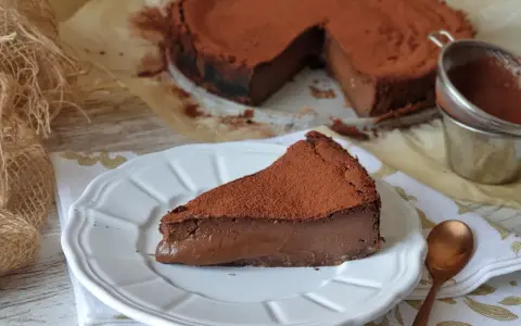 Una rebanada de torta de chocolate, cubierta de cacao en polvo, reposa en un plato blanco. El postre se ve cremoso y delicioso.