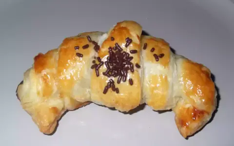 Croissant de chocolate recién horneado, dorado y brillante, espolvoreado con virutas de chocolate.