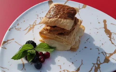 Delicioso postre de milhojas de crema, con capas crujientes y relleno dulce, acompañado de frutos rojos y menta fresca.