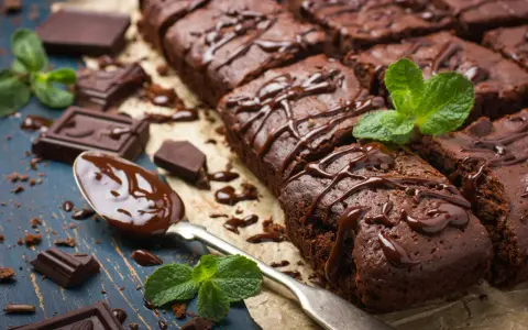 Deliciosos postres de chocolate, brownies cubiertos con glaseado de chocolate y decorados con menta fresca. Trozos de chocolate y una cuchara con chocolate derretido completan la escena.