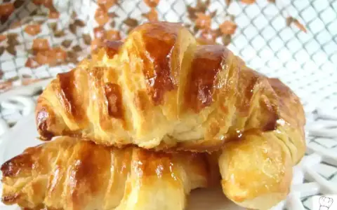 Dos croissants caseros dorados y brillantes, apilados en un plato blanco.