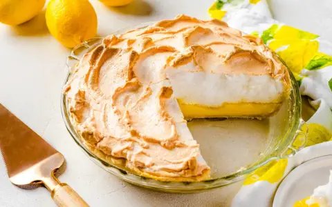 Tarta de limón con merengue dorado y esponjoso, lista para servir. Dos limones frescos y un paño con estampado de limones completan la escena.