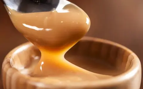 Cuchara vertiendo manjar blanco cremoso en un cuenco de madera.