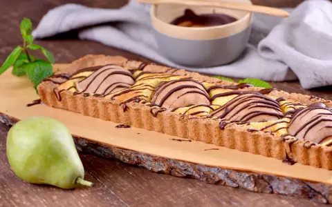 Tarta rectangular con peras y chocolate, decorada con menta. Una pera fresca reposa a un lado.