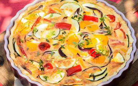 Quiche de verduras con hojaldre, repleta de pimientos, calabacín y cebolla morada, adornada con hierbas frescas.