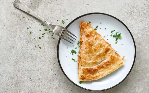 Una porción dorada de empanada de atún, lista para ser disfrutada con un tenedor plateado y espolvoreada con perejil fresco.