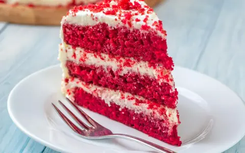 Rebanada de torta red velvet con glaseado blanco y migas rojas, servida en plato blanco con tenedor.