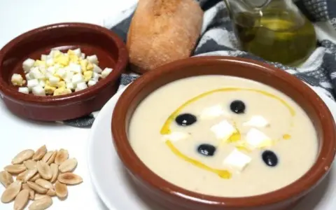 Un cuenco de mazamorra cordobesa con aceitunas, huevo duro y un chorrito de aceite. Acompaña un panecillo y almendras.