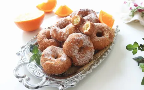 Rosquillas de naranja recién hechas, espolvoreadas con azúcar, junto a gajos de naranja fresca en una bandeja plateada.