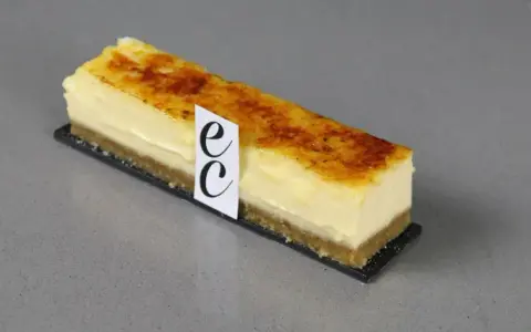 Un trozo de cheesecake creme brulee con base crujiente y cubierta caramelizada, adornado con un distintivo.
