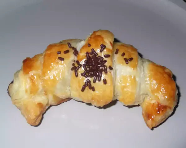 Croissant de chocolate recién horneado, dorado y brillante, espolvoreado con virutas de chocolate.