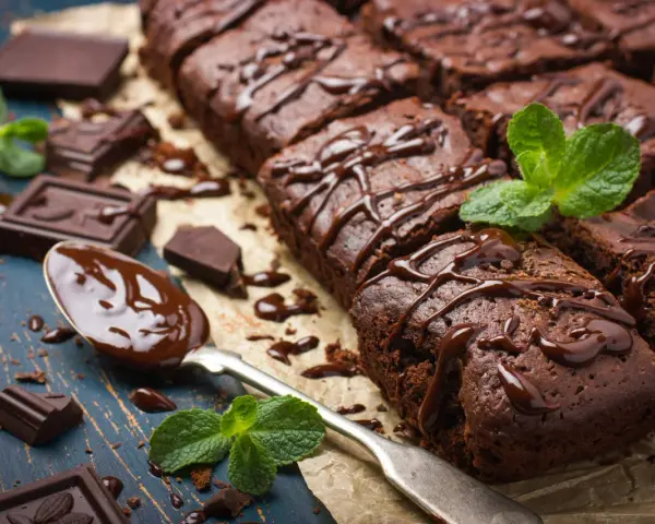 Deliciosos postres de chocolate, brownies cubiertos con glaseado de chocolate y decorados con menta fresca. Trozos de chocolate y una cuchara con chocolate derretido completan la escena.