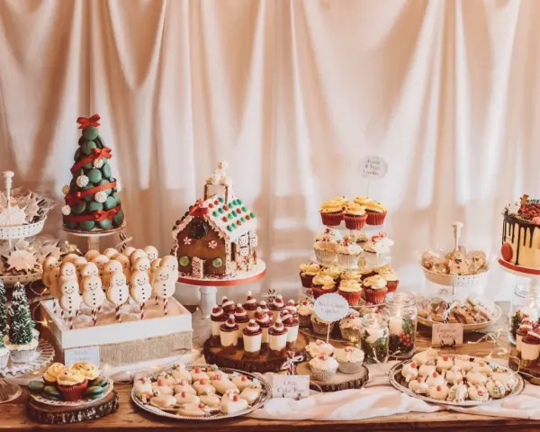 Mesa llena de postres navideños: árbol de macarons, casa de jengibre, muñecos de nieve, cupcakes y galletas.