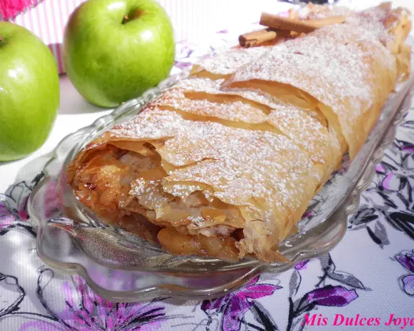 Delicioso strudel de manzana recién horneado, espolvoreado con azúcar glas y acompañado de manzanas verdes frescas y canela.