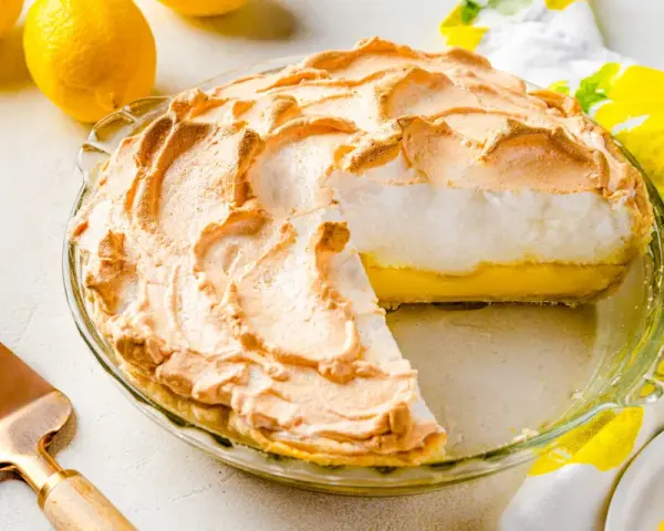 Tarta de limón con merengue dorado y esponjoso, lista para servir. Dos limones frescos y un paño con estampado de limones completan la escena.