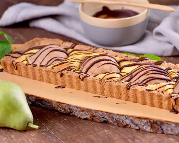 Tarta rectangular con peras y chocolate, decorada con menta. Una pera fresca reposa a un lado.