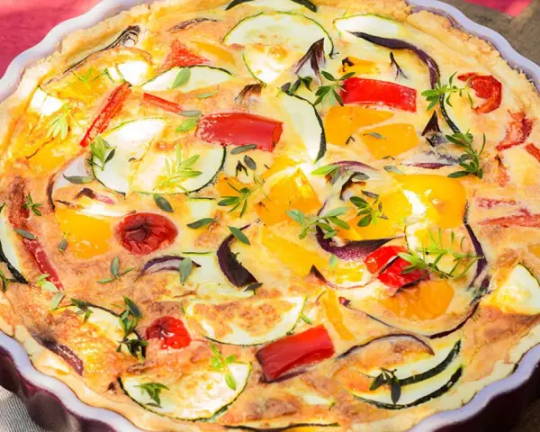 Quiche de verduras con hojaldre, repleta de pimientos, calabacín y cebolla morada, adornada con hierbas frescas.