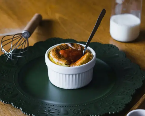 Un esponjoso soufflé con salsa de tomate, listo para disfrutar. Al lado, un batidor y un vaso de leche, ingredientes clave para crear que es un soufflé.