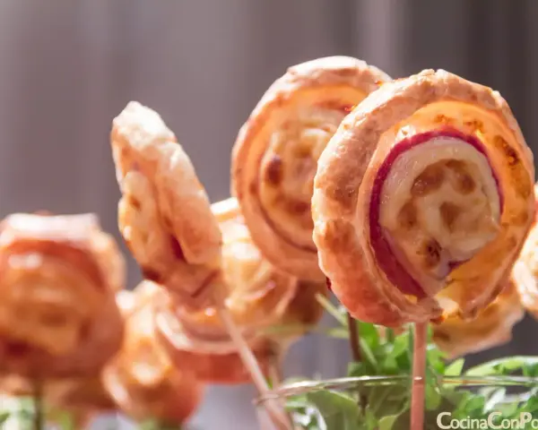 Deliciosas piruletas de hojaldre rellenas de queso y jamón, presentadas en un vaso con hojas verdes.